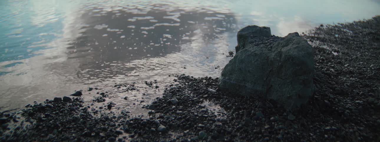 A tranquil close-up of calm water gently lapping against a rocky, pebbled shore at dusk