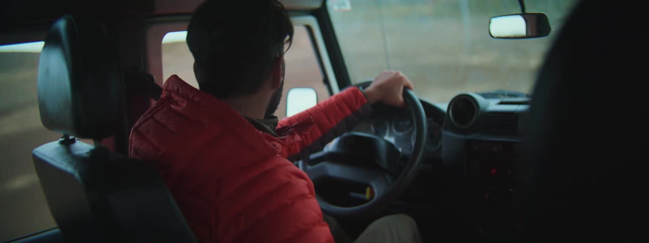 A focused man with a beard wearing a red puffer jacket drives his rugged vehicle on a country road