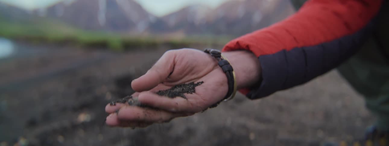 Close-up of a person's hand as dark volcanic sand slowly slips through their fingers with mountains in the background