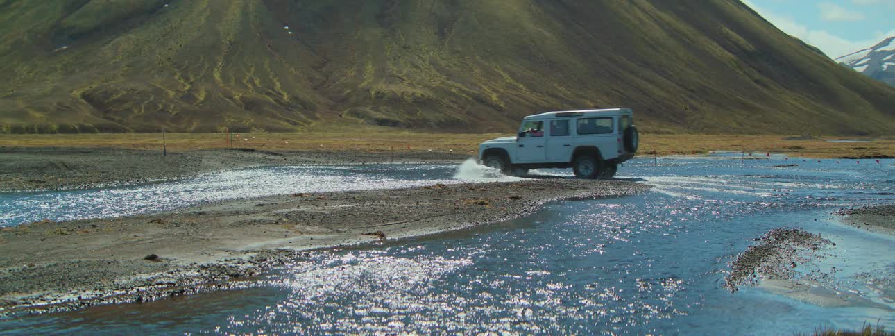 A white 4x4 fords a sparkling river in slow motion through a remote mountain landscape