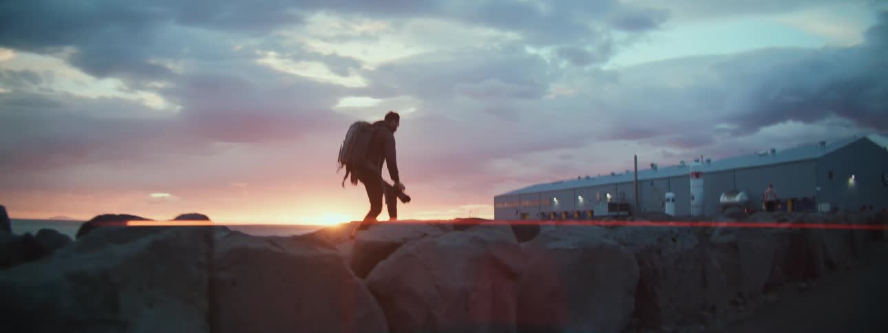 A silhouetted adventurer with a backpack and camera carefully walks across a rocky jetty at sunset