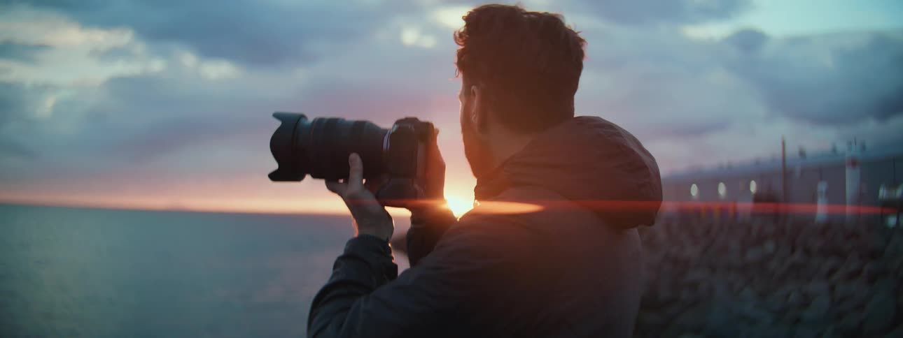 A professional photographer in a jacket captures the beautiful coastal sunset over the ocean with his camera