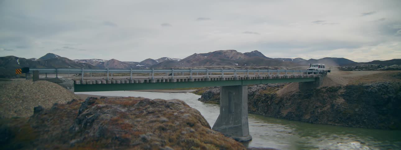 A white SUV drives over a bridge in the remote, rugged landscape of the Icelandic highlands