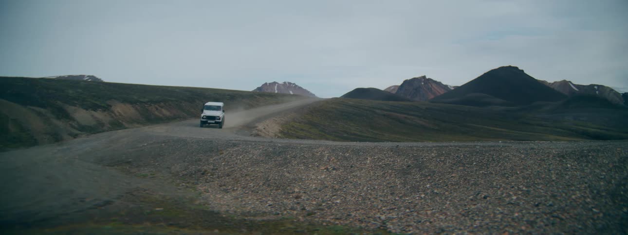 A white 4x4 SUV navigates a rugged, winding gravel road before crossing a bridge over a river in a vast, remote landscape
