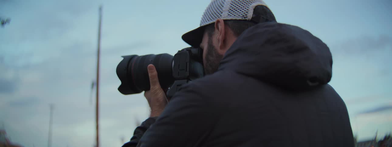 A professional photographer with a beard looks through the viewfinder of his digital camera outdoors