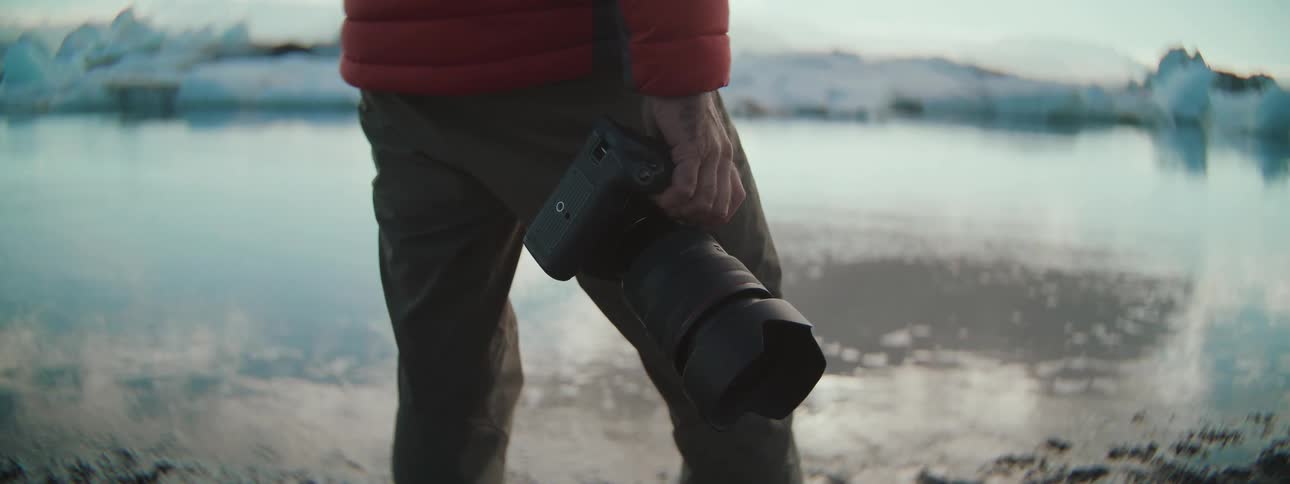 Close-up of a photographer in a red jacket holding a professional camera while standing by a glacial lagoon