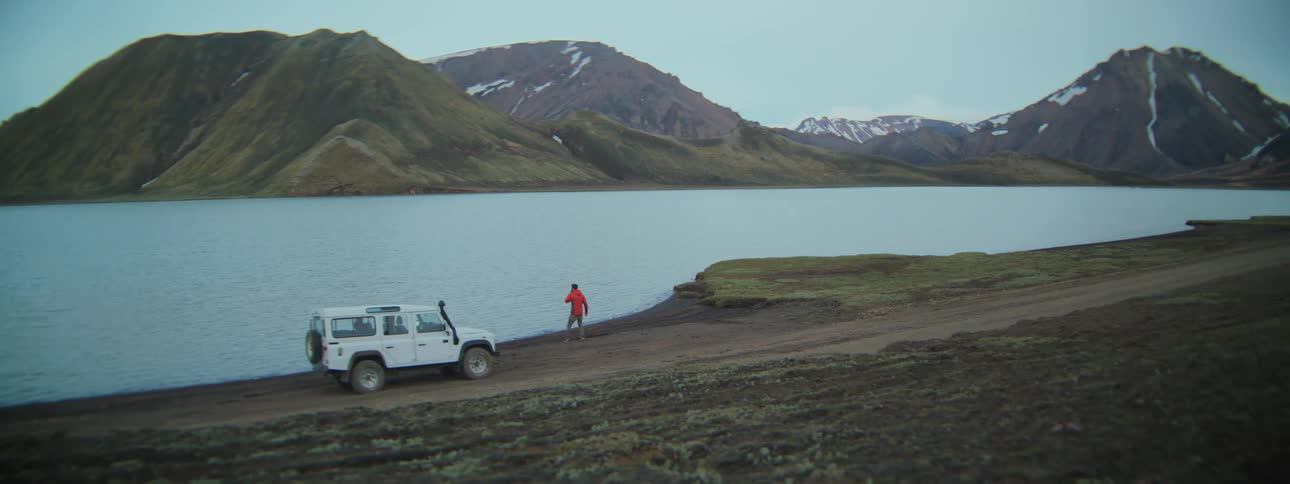 A traveler in a red jacket gets out of their white 4x4 vehicle to admire a tranquil lake in a remote, mountainous wilderness