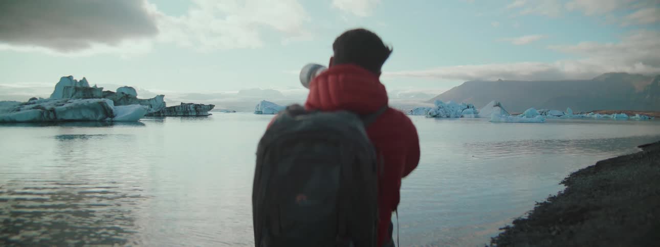 A photographer wearing a red jacket and backpack captures images of majestic icebergs floating in a glacial lagoon