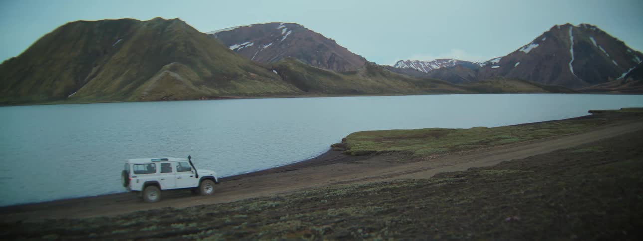 A white off-road SUV drives up and parks on the shore of a tranquil lake surrounded by stunning, snow-dusted mountains
