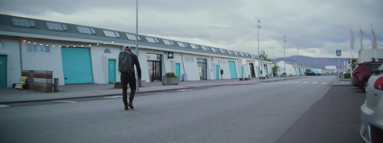 A lone traveler with a large backpack walks away from the camera down a quiet street next to a row of modern shops