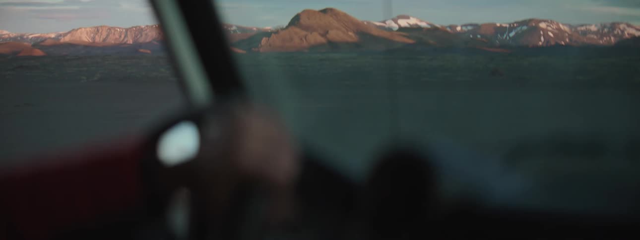 An over-the-shoulder perspective from a moving car captures the sunlit, snow-capped mountains of a vast, rugged landscape