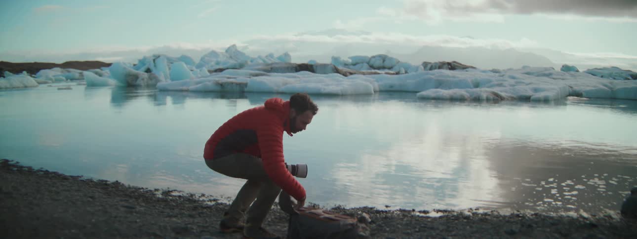 A male photographer in a red jacket takes off his backpack to set up his camera by a stunning glacier lagoon