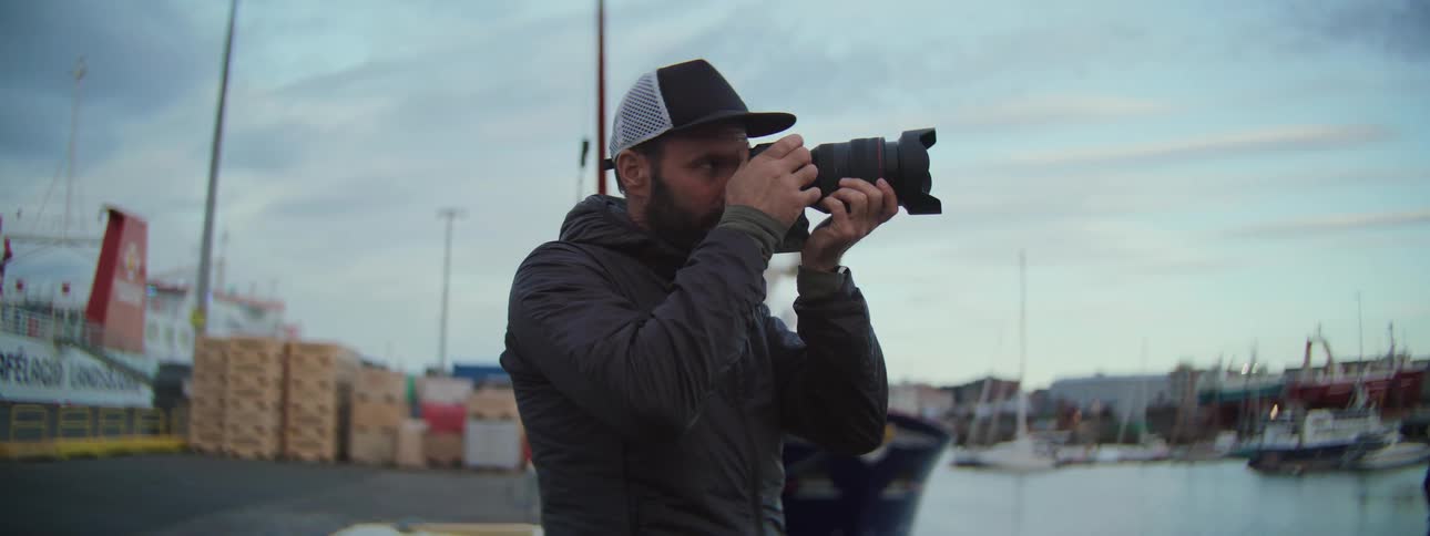 A bearded photographer in a cap focuses his camera lens while standing at a bustling fishing harbor at dusk