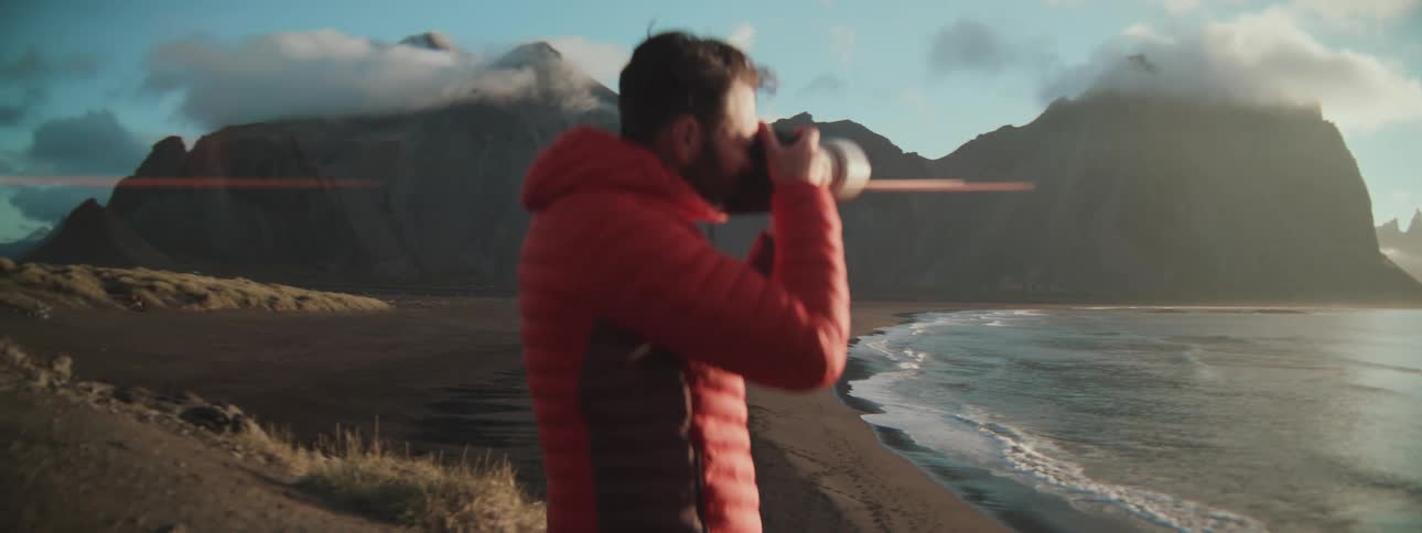A professional photographer in a red jacket captures the stunning scenery of a black sand beach and mountains at sunrise