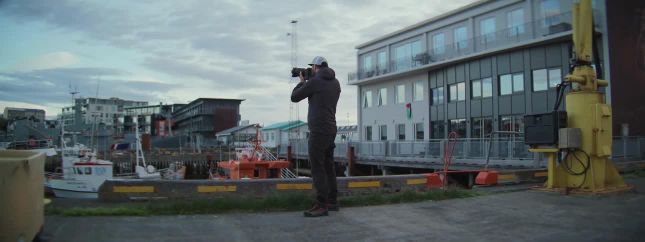A photographer stands on a dock in a coastal town, taking pictures of the harbor on an overcast day