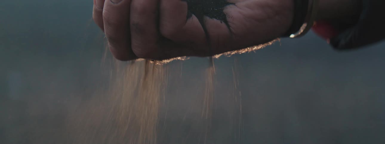 Close-up slow-motion shot of a dirty hand letting dark sand fall through its fingers with the sunset backlighting the scene
