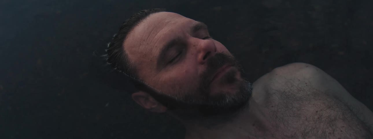 A close-up of a bearded man's face as he floats serenely in dark water, slowly opening his eyes