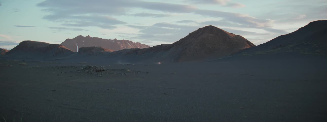 A lone vehicle with its headlights on drives across a vast, barren landscape at twilight with mountains in the distance