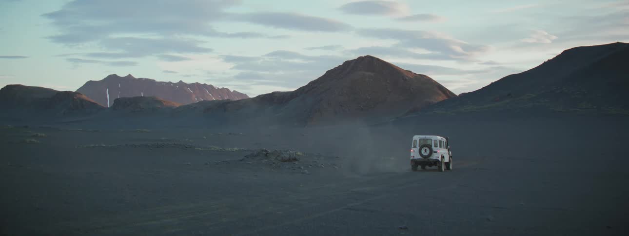 A white off-road vehicle drives through a vast and desolate volcanic landscape towards mountains in the distance at dusk