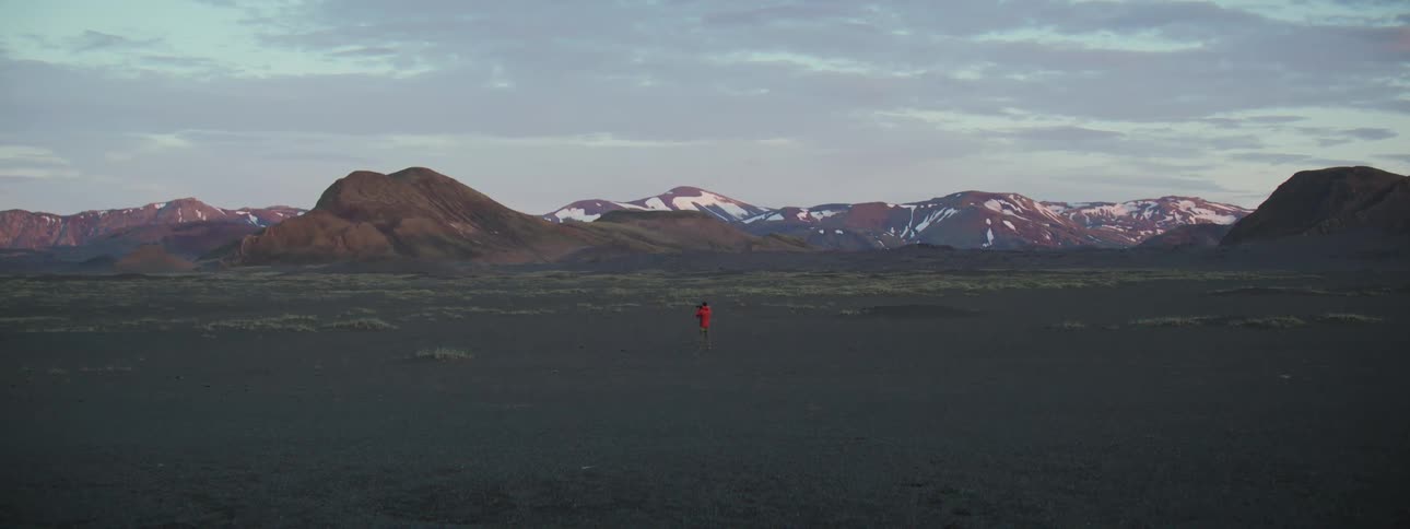 A lone photographer in a red jacket stands in a desolate volcanic landscape, capturing images of the snow-capped mountains at dusk