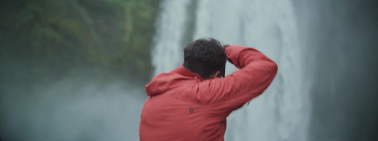 A photographer wearing a bright red jacket takes pictures of a majestic, misty waterfall in slow motion