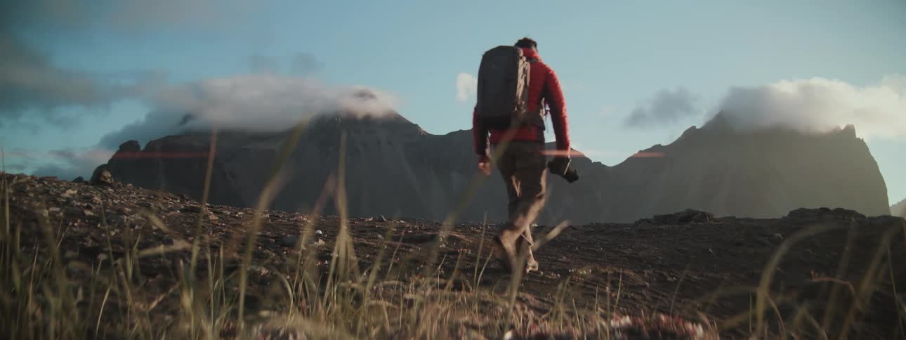A male photographer with a backpack walks through a vast, rocky landscape towards cloud-covered mountains in slow motion