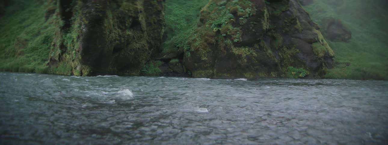 Rain falls on a flowing river in a lush, green, mossy canyon