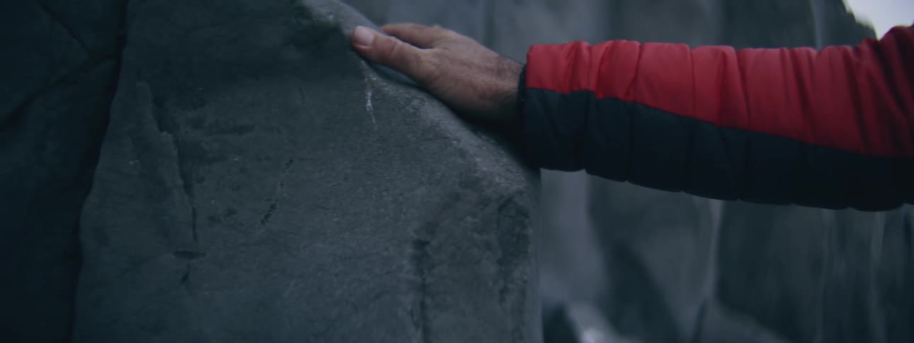 A person's hand wearing a red puffer jacket sleeve rests on a large, cool-toned rock in nature