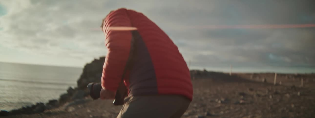 A male photographer in a red jacket walks along a rugged, sun-kissed coastline and prepares to capture the stunning landscape