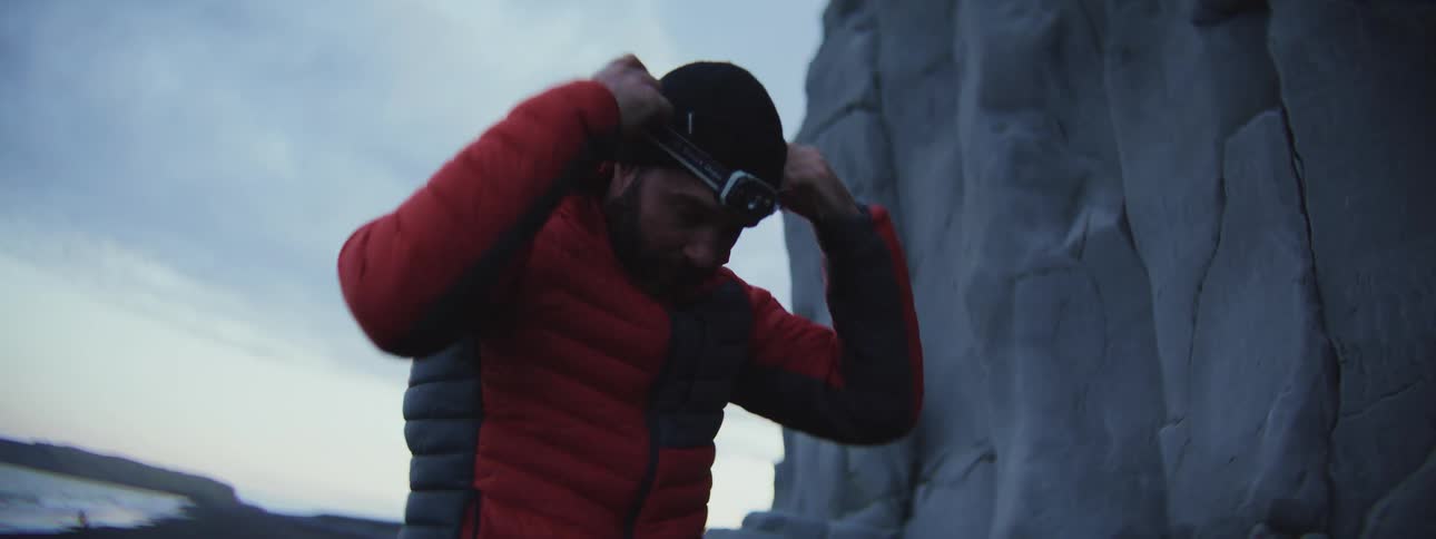 A man in a red jacket and beanie prepares for a night climb by putting on his headlamp next to a rock formation