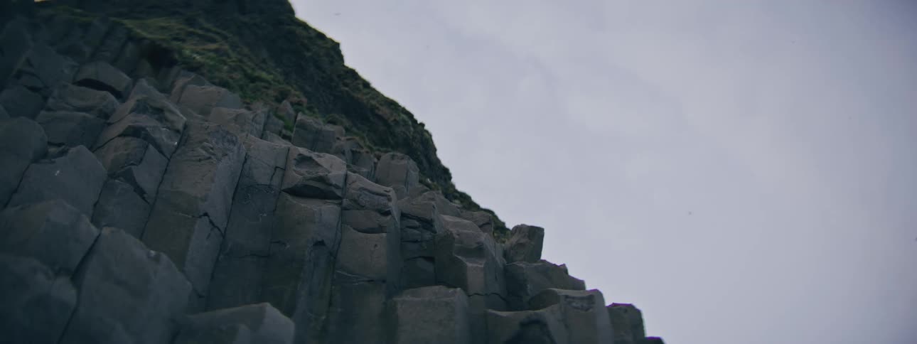 A low-angle view of rugged basalt column cliffs topped with green moss under an overcast sky