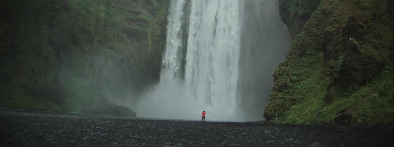 A tiny figure in a red jacket is dwarfed by the immense power of a massive waterfall cascading down a lush green cliff in Iceland