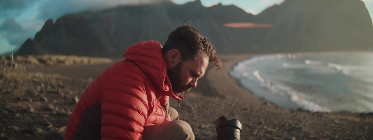 A photographer crouches on a bluff overlooking the ocean and mountains as he assembles his professional camera and lens