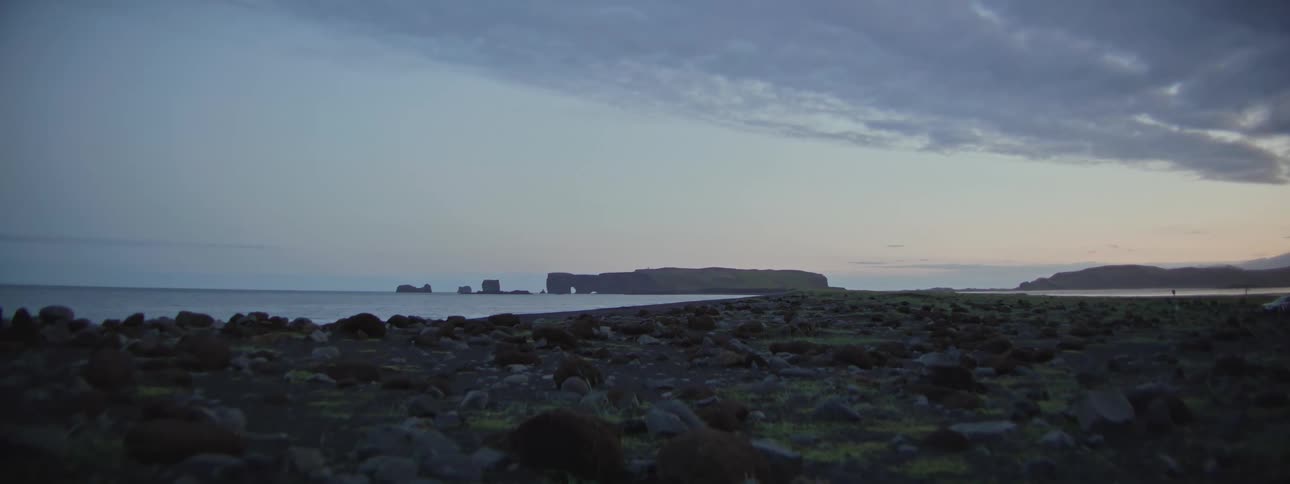 A serene, wide shot shows the Dyrhólaey peninsula and its flashing lighthouse from a rocky beach at dusk in Iceland