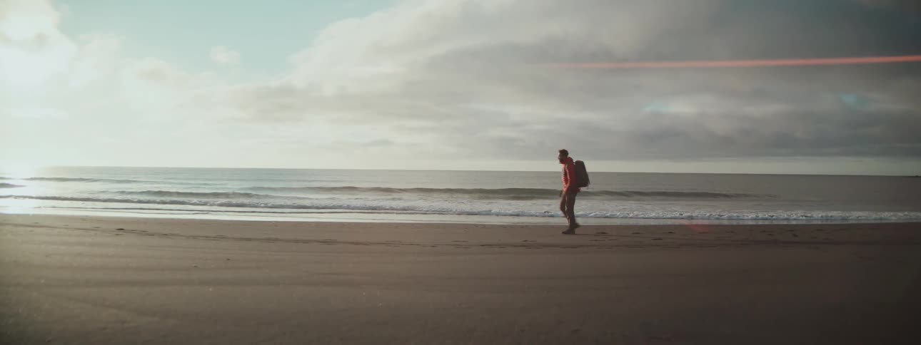 A lone hiker with a backpack walks along the sandy shoreline and stops to look out at the ocean waves