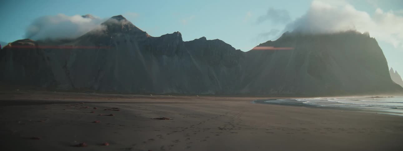 A serene and vast black sand beach stretches out before a majestic, cloud-capped mountain range at sunrise