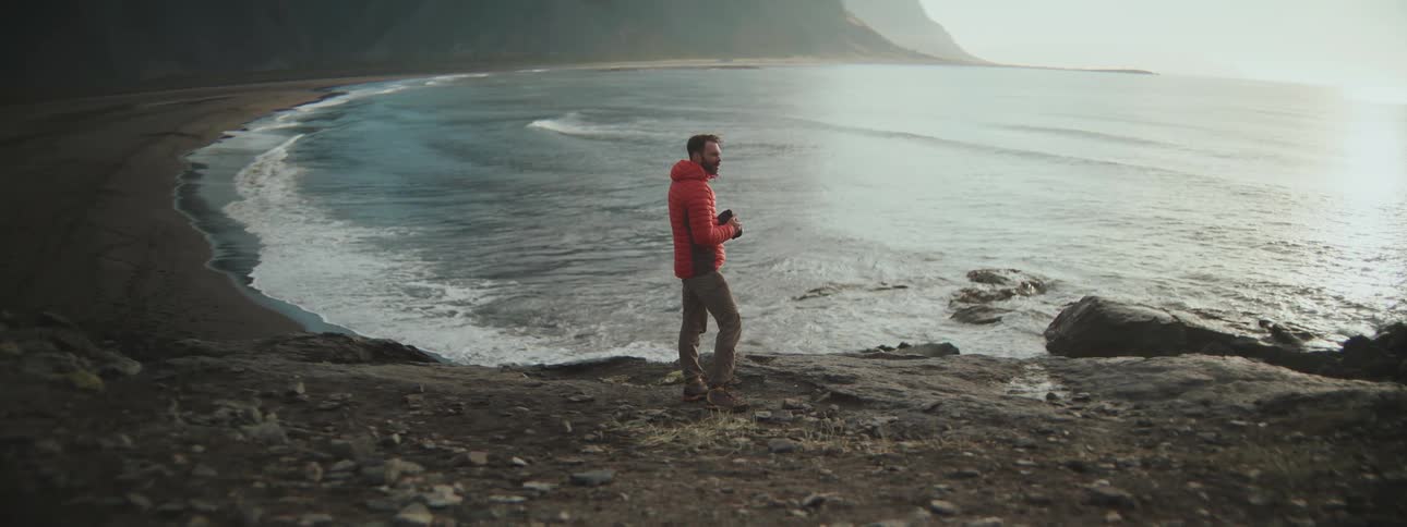 A man in a red jacket stands on a rocky cliff overlooking a vast ocean bay, taking a picture of the scenic landscape