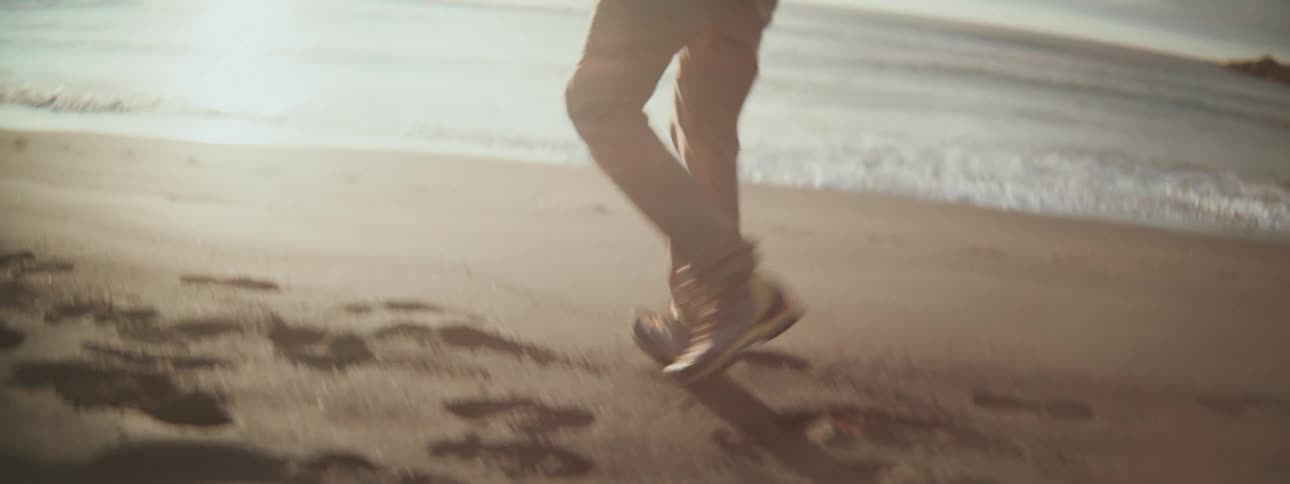 A photographer with a backpack hikes along a sandy beach, leaving footprints as he stops to capture the ocean at sunset