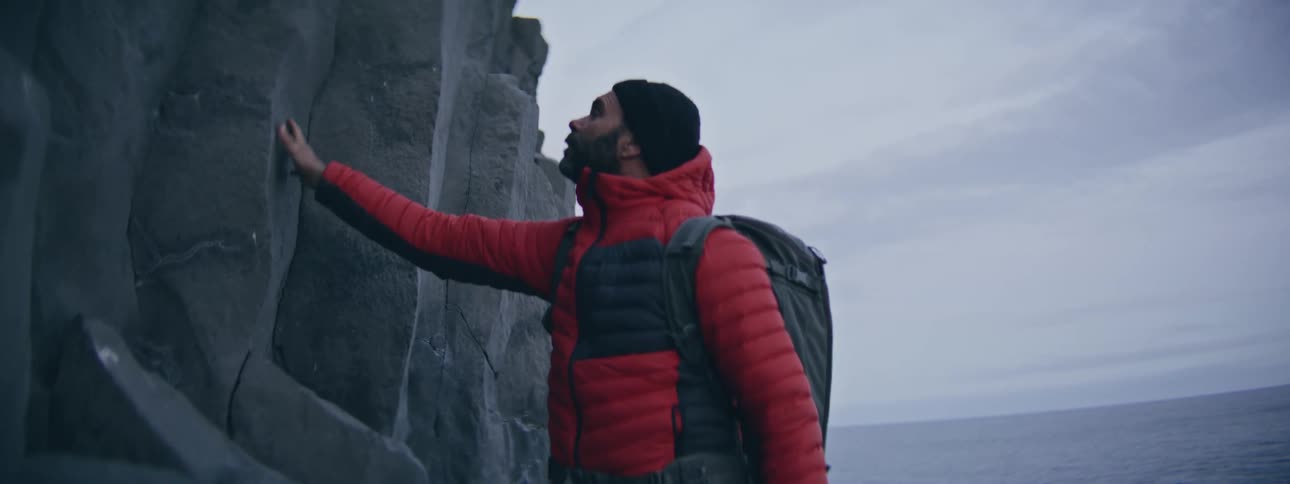 An adventurous man in a red jacket and backpack stands in awe, touching a giant coastal rock formation