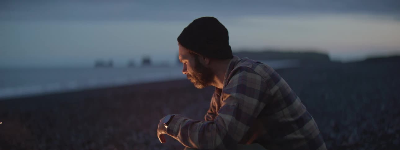 A thoughtful man wearing a beanie and plaid shirt sits by a beach campfire at dusk, gazing into the flames