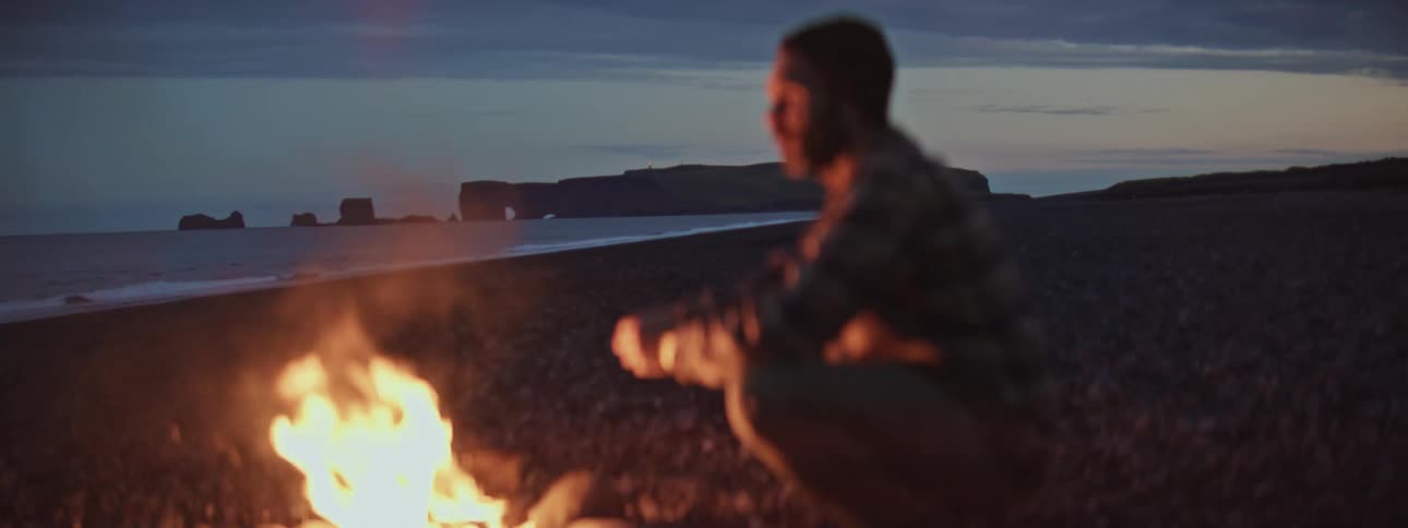 An out-of-focus person warms themselves by a campfire on a rocky beach at dusk, with dramatic coastal cliffs in the distance