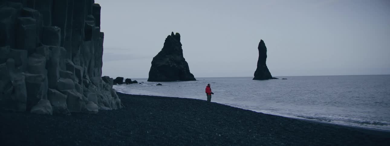 A lone person in a red jacket stands on a desolate black sand beach, gazing at dramatic sea stacks rising from the ocean