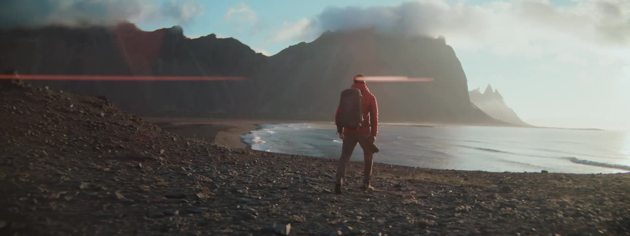 A photographer with a backpack hikes down a rocky path towards a breathtaking Icelandic coastline with mountains and the ocean at sunrise
