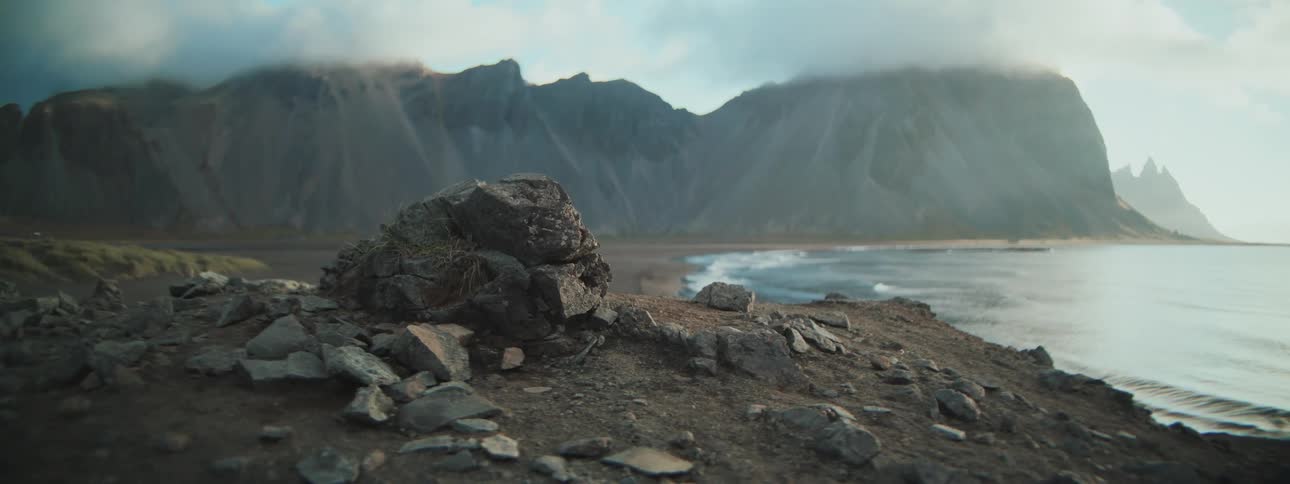 A scenic landscape shot from a rocky cliff reveals the ocean waves washing up on a dark sand beach with a vast, cloudy mountain range in the distance