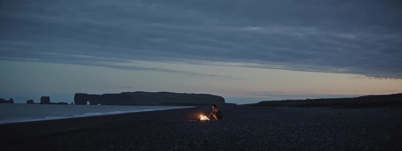 A solitary person sits by a glowing campfire on a vast and remote black sand beach at dusk