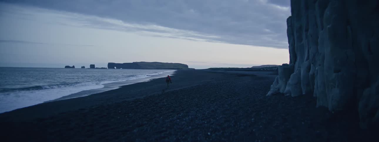 A solitary hiker in a red jacket walks along the moody and dramatic black sand beach of Reynisfjara in Iceland