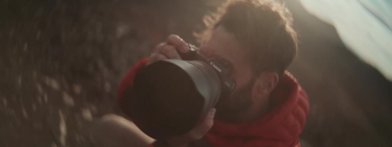 A male photographer in a red jacket focuses his professional camera with a telephoto lens, captured in a slow-motion rack focus with a warm lens flare
