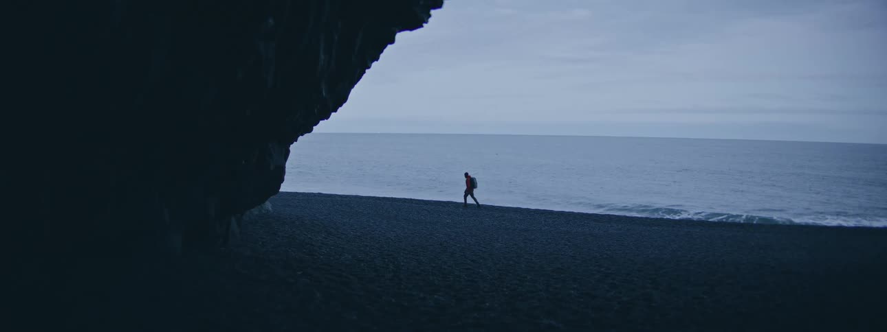 A lone hiker in a red jacket walks along the shoreline of a vast and moody black sand beach