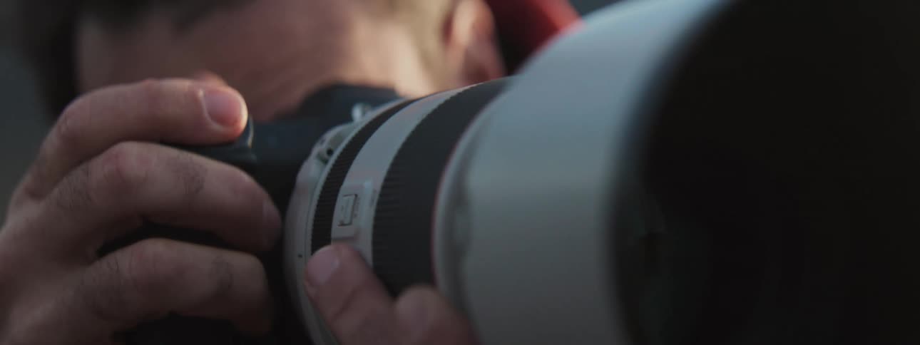 A slow motion close-up of a photographer's hands as they hold and operate a professional camera with a large telephoto lens