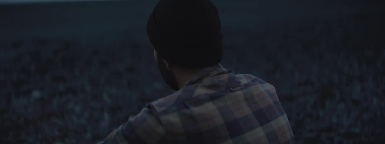 A bearded man in a beanie and plaid shirt warms himself by a campfire on a dark beach at twilight
